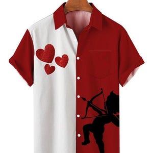 May include: A short-sleeved button-down shirt with a split design. One side is white with three red heart graphics, and the other is red with a black silhouette of Cupid aiming a bow and arrow. The shirt is hanging on a wooden hanger.