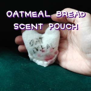 May include: A heart-shaped, white scent pouch with the words "OATMEAL BREAD SCENT POUCH" in purple and white text. The pouch has the words "OAT MEAL" written on it in black and red ink. The pouch is held in a hand.