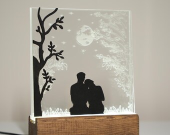 Könnte beinhalten: Beleuchtete Glasscheibe mit einer Silhouette eines Paares unter einem Baum, Mond und Sternen. Die schwarzen Silhouetten heben sich von einem mattierten Hintergrund ab. Das Panel steht auf einem rechteckigen Holzsockel.
