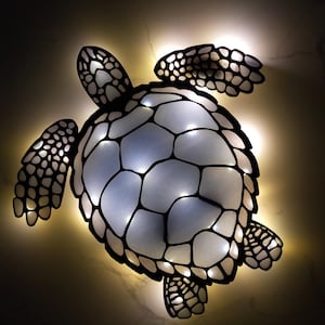 Puede incluir: Una lámpara decorativa de pared con forma de tortuga marina. La tortuga es negra con un caparazón blanco y azul claro, iluminado con pequeñas luces. Las aletas y la cabeza también son negras. La lámpara está diseñada para montarse en la pared.