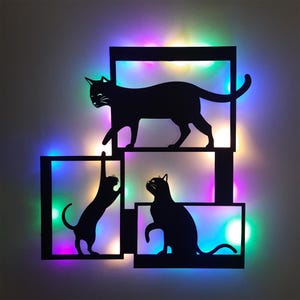 Könnte beinhalten: Schwarze Metall-Wandkunst mit drei Katzen-Silhouetten in rechteckigen Rahmen. Die Katzen sind in verschiedenen Posen, mit farbigen LED-Lichtern, die den Hintergrund beleuchten. Das Kunstwerk ist für die Wandmontage konzipiert.
