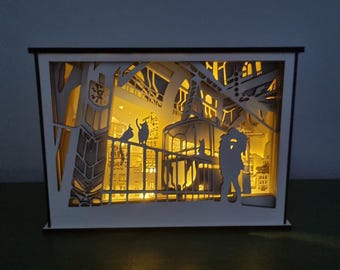 May include: A layered paper art light box featuring a cityscape scene with cats on a balcony and a couple embracing. The artwork is illuminated from within, creating a warm, inviting glow. The box has a black frame and a white interior.