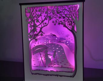 Könnte beinhalten: Ein geschichteter Papierkunst-Leuchtkasten mit einer malerischen Außenlandschaft, die eine Person unter einem Regenschirm beim Malen zeigt. Das Kunstwerk wird von einem sanften lilafarbenen Schein beleuchtet, der die Bäume, die Straße und die Häuser hervorhebt. Der Rahmen ist weiß mit einem schwarzen Rand.