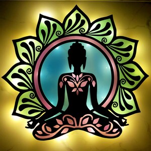 May include: A black silhouette of a meditating Buddha figure, centered within a lotus flower design. The lotus petals are green with black detailing, surrounding a pink and blue circular background. The image is illuminated with a warm, yellow glow.