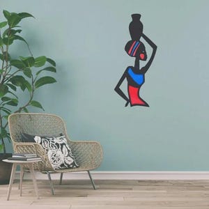 May include: A wall art piece depicting a stylized figure of a woman carrying a vessel on her head. The figure is black with red, blue, and striped accents. The artwork is mounted on a light blue wall, with a chair and plant in the foreground.