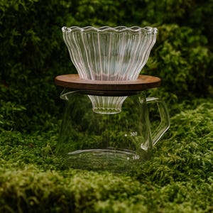 Könnte beinhalten: Ein klares Kaffee-Pour-Over-Set aus Glas mit Holzsockel und Glaskaraffe. Die Karaffe hat einen Griff und Messmarkierungen für 150 und 300 ml. Der Pour-Over hat ein geripptes Design.