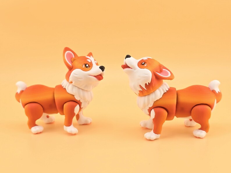 Pu&ograve; includere: Due statuette di cani corgi arancioni e bianchi con giunture articolate. Un cane guarda in alto con la bocca aperta, l'altro guarda avanti con la lingua di fuori. Le statuette hanno pelliccia bianca intorno al collo.