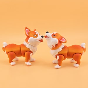 May include: Two orange and white corgi dog figurines. One dog is looking at the other with its tongue out. The other dog is looking up with its mouth open. The figurines have articulated joints.