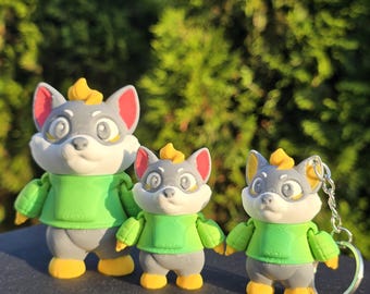 May include: Three cartoon fox figurines. The foxes have gray faces, white chests, and green shirts. They have yellow accents on their heads and feet. One fox is a keychain. The background is blurred green foliage.