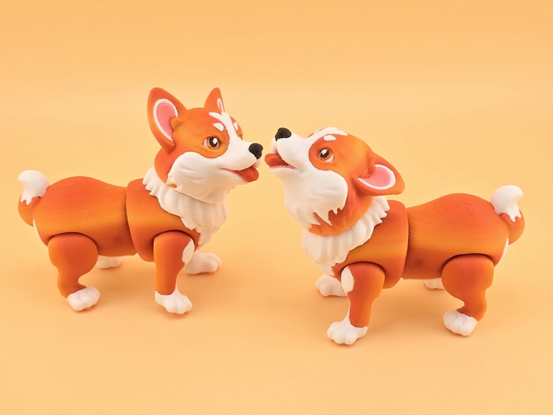 Pu&ograve; includere: Due statuette di cani corgi arancioni e bianchi. I cani hanno la pelliccia bianca intorno al collo e sulle zampe. Un cane guarda in alto con la lingua di fuori, e l'altro lo guarda. Lo sfondo &egrave; arancione chiaro uniforme.