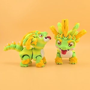 Könnte beinhalten: Zwei skurrile Drachenfiguren in Grün und Gelb mit ausdrucksstarken Gesichtern und verspielten Posen. Ein Drache steht, der andere geht. Die Figuren haben gelbe Akzente an Füßen, Mähnen und Hörnern, vor einem einfarbigen orangefarbenen Hintergrund.