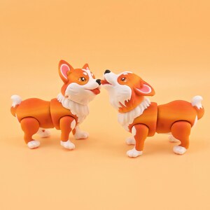 Pu&ograve; includere: Due statuette di cani corgi arancioni e bianchi. I corgi sono in posa giocosa, uno lecca la faccia dell'altro. Le statuette hanno giunture articolate e pelliccia bianca intorno al collo.