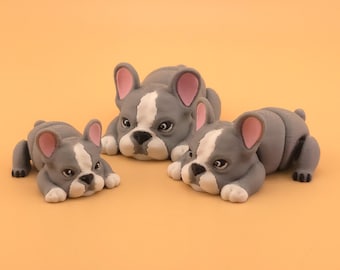 May include: Three gray and white bulldog figurines with pink ears and black paws are arranged on an orange background. The bulldogs are lying down, with their heads and paws visible. The figurines vary in size, suggesting a family set.