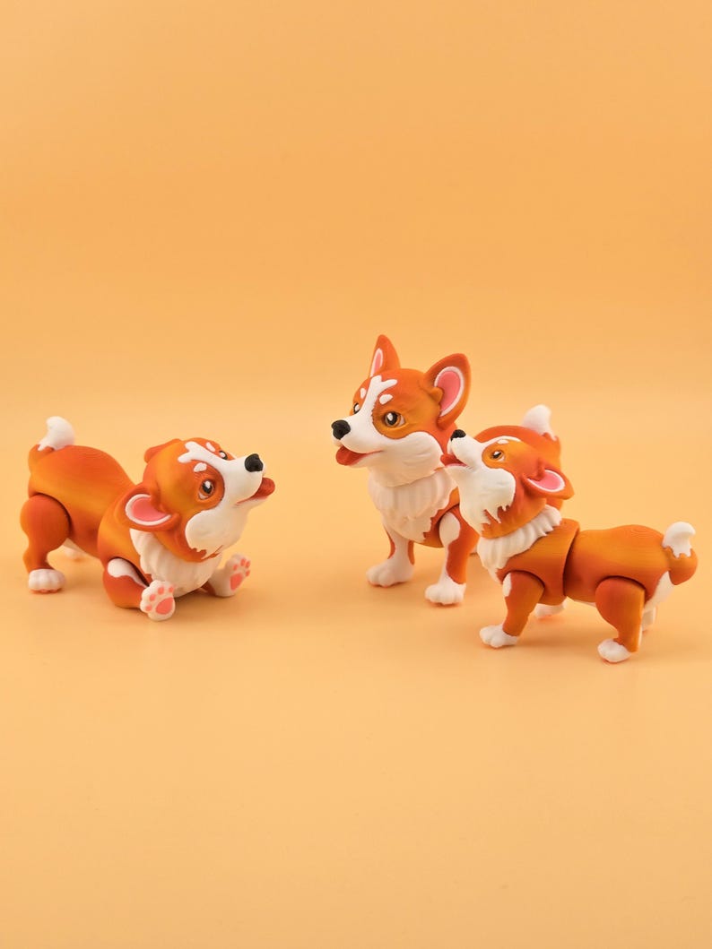 Pu&ograve; includere: Tre figurine di corgi arancioni e bianchi sono esposte su uno sfondo arancione uniforme. Una statuina &egrave; sdraiata, un'altra &egrave; in piedi e la terza guarda in alto. I corgi hanno giunture articolate.