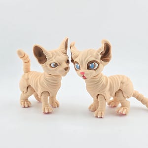 May include: Two articulated beige cat figurines with wrinkled skin textures, large pointed ears, and blue eyes. The figures feature segmented tails and limbs, with one cat standing upright and the other positioned with its head turned slightly forward.