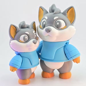 Op de afbeelding: Twee 3D-geprinte wolfsbeeldjes. De wolven zijn grijs met witte en oranje accenten, gekleed in blauwe jassen. De grotere wolf staat naast de kleinere. De beeldjes hebben een cartoonachtig ontwerp en staan tegen een witte achtergrond.