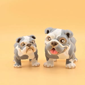 May include: Two bulldog figurines, one large and one small, stand side-by-side against a yellow background. The bulldogs are gray and white with detailed facial features, including open mouths and pink tongues. The figurines are likely decorative.