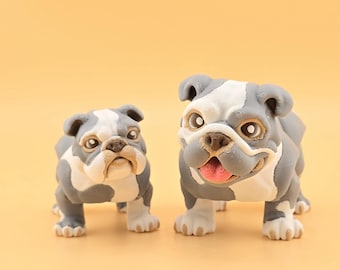 Può includere: Due statuette di bulldog, una grande e una piccola, sono affiancate su uno sfondo giallo. I bulldog sono grigi e bianchi con dettagli del viso, tra cui bocche aperte e lingue rosa. Statuette decorative.