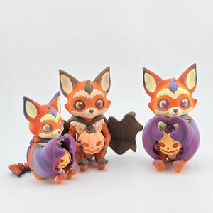 May include: Three orange and brown fox figurines with large ears and wide eyes. Each fox holds a small pumpkin. Two foxes have purple bat-like wings. The figurines are set against a white background.
