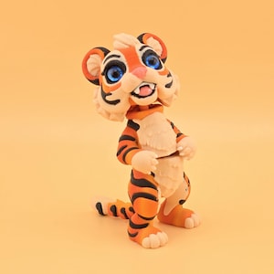 Op de afbeelding: Een handgemaakt tijgerbeeldje met oranje en zwarte strepen, blauwe ogen en een crèmekleurige borst. De tijger staat in een speelse pose, met zijn voorpoten omhoog en één knie gebogen. Het beeldje staat tegen een effen oranje achtergrond.