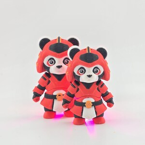 Op de afbeelding: Twee panda beeldjes in rode samoerai-pantser met zwarte accenten. De panda's hebben witte gezichten, zwarte oren en rode helmen. Het pantser omvat een rok en borststuk. De beeldjes staan op een witte ondergrond.