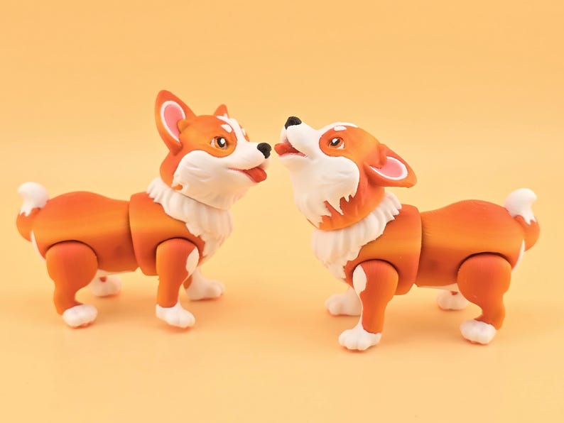 Pu&ograve; includere: Due statuette di cani corgi arancioni e bianchi. I cani sono uno di fronte all'altro, uno con la lingua di fuori. Le statuette hanno giunture articolate e pelliccia bianca intorno al collo e sulle zampe.