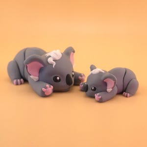 May include: Two gray koala figurines, one larger and one smaller, are lying down on an orange surface. The koalas have pink ears and feet, black noses, and white accents on their heads. The figurines are detailed and appear handmade.