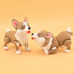 Pu&ograve; includere: Due statuette di cani corgi marroni e bianchi con dettagli rosa. Uno &egrave; in piedi, l'altro &egrave; in posa giocosa. Le statuette hanno pelliccia bianca intorno al collo, su sfondo giallo.