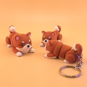 May include: Three Shiba Inu dog figurines in shades of brown, white, and pink. One is attached to a silver keychain. The dogs are in a lying down position. The background is a solid orange color.
