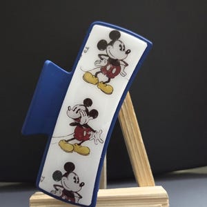 Könnte beinhalten: Eine blaue Haarspange mit einem weißen, rechteckigen Mittelteil, das mit drei sich wiederholenden Illustrationen von Mickey Mouse verziert ist. Jede Figur ist im klassischen Stil mit roter Hose und gelben Schuhen dargestellt und vertikal angeordnet.