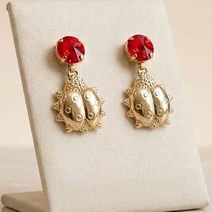 May include: A pair of gold-toned drop earrings featuring a ladybug design. Each earring has a round, faceted red crystal at the top and a textured gold-colored metal ladybug charm suspended below, showcasing detailed wings and small circular markings.