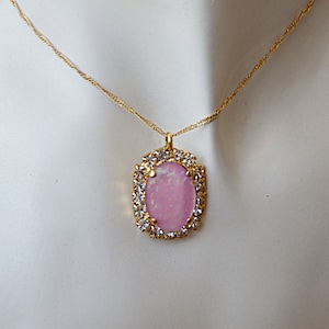 Pu&ograve; includere: Una collana con catena dorata con un ciondolo ovale rosa incastonato in una cornice dorata con strass trasparenti.