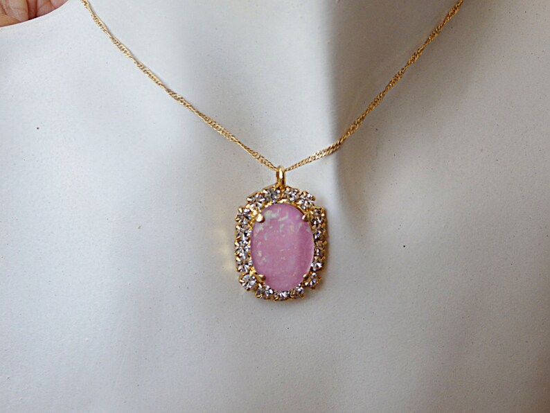 Pu&ograve; includere: Una collana con catena dorata con un ciondolo ovale rosa incastonato in una cornice dorata con strass trasparenti.