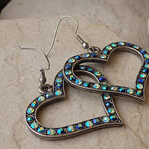 Pu&ograve; includere: Un paio di orecchini a forma di cuore argentati con strass blu e verdi iridescenti.