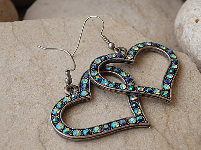 Pu&ograve; includere: Un paio di orecchini a forma di cuore argentati con strass blu e verdi iridescenti.