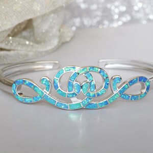 May include: A silver bracelet with a blue opal design. The design features two intertwined circles with a swirling pattern.