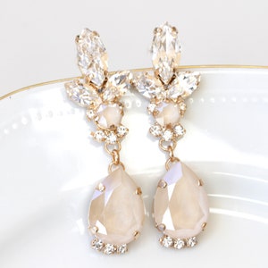 May include: A pair of gold tone dangle earrings with clear and iridescent white crystals. The earrings feature a teardrop shaped crystal at the bottom.