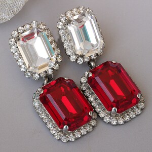 May include: A pair of dangle earrings with a clear crystal stone and a red crystal stone on each earring. The stones are surrounded by a silver setting with smaller clear crystals.
