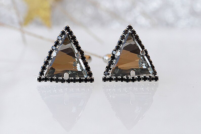 Puede incluir: Un par de pendientes de forma triangular con una piedra preciosa gris transparente engastada en un marco de plata. El marco est&aacute; acentuado con una fila de piedras preciosas negras.