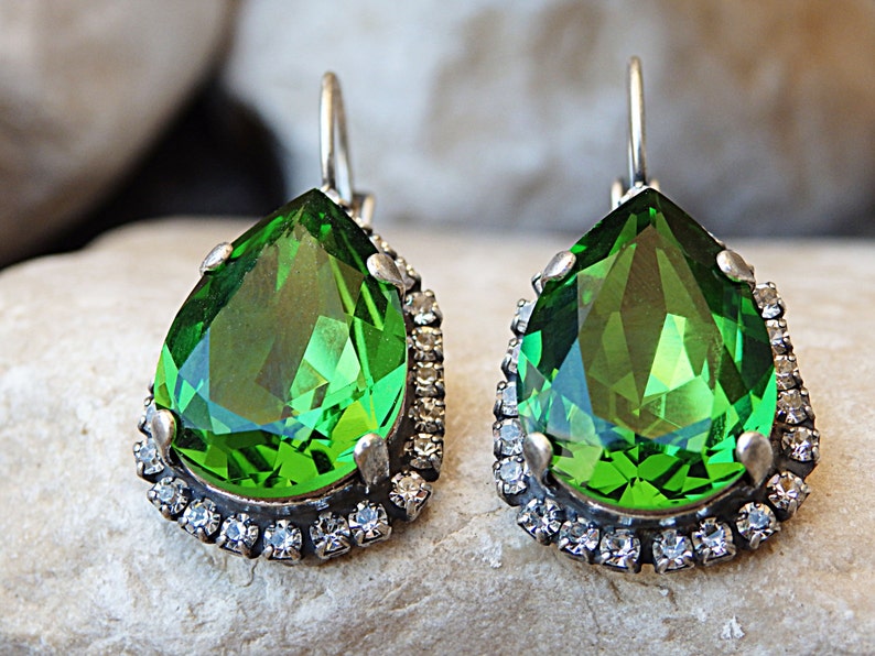 May include: A pair of dangle earrings with a large pear-shaped green gemstone surrounded by a halo of smaller clear rhinestones.