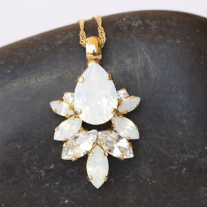 May include: A gold pendant necklace with a large, pear-shaped, opalescent crystal at its centre, encircled by smaller, clear crystals. The pendant is suspended from a delicate gold chain. The necklace is displayed on a dark, textured surface.