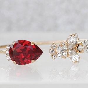 May include: A gold bangle bracelet with a large red teardrop-shaped crystal on one end and a cluster of smaller clear crystals on the other end.