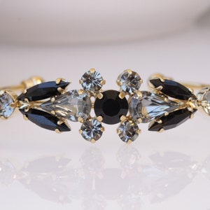 Puede incluir: Un brazalete de oro con diamantes de imitación negros y transparentes. El brazalete está adornado con un grupo de piedras negras y transparentes en el centro.