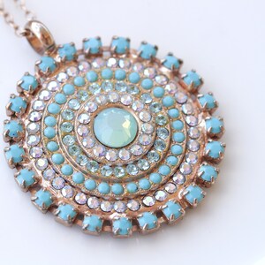 May include: A gold pendant necklace with a round turquoise and clear crystal design. The pendant features a large, light blue center stone surrounded by concentric circles of smaller turquoise and clear crystals.