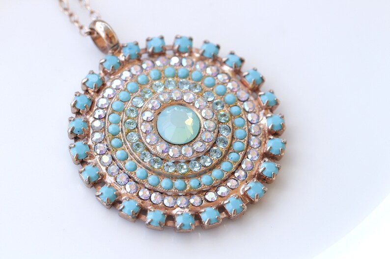 May include: A gold pendant necklace with a round turquoise and clear crystal design. The pendant features a large, light blue center stone surrounded by concentric circles of smaller turquoise and clear crystals.