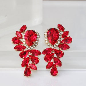May include: A pair of dangle earrings with a teardrop-shaped red crystal surrounded by smaller red crystals and clear rhinestones.