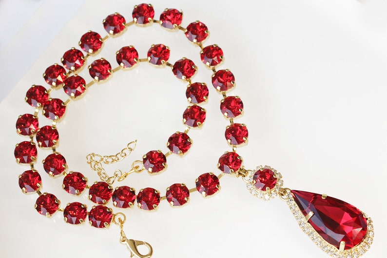 May include: A red crystal necklace with a teardrop pendant. The necklace is made of small, round crystals that are connected by gold-colored metal.