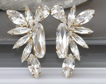 May include: A pair of gold tone earrings with clear crystal embellishments. The earrings are shaped like a fan and have a large, clear crystal in the center.
