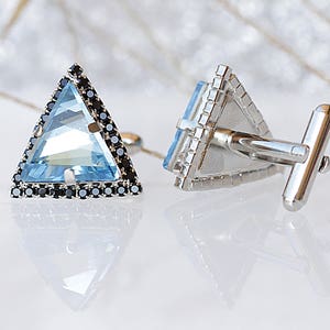 Puede incluir: Un par de gemelos de plata con una piedra preciosa azul triangular rodeada de piedras negras. Los gemelos tienen una barra de plata para conectarlos a la camisa.