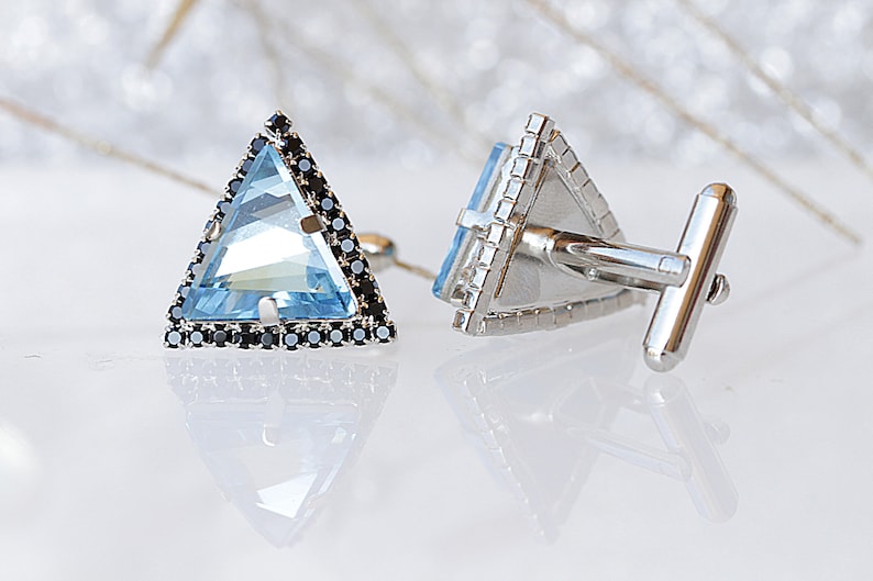 Puede incluir: Un par de gemelos de plata con una piedra preciosa azul triangular rodeada de piedras negras. Los gemelos tienen una barra de plata para conectarlos a la camisa.